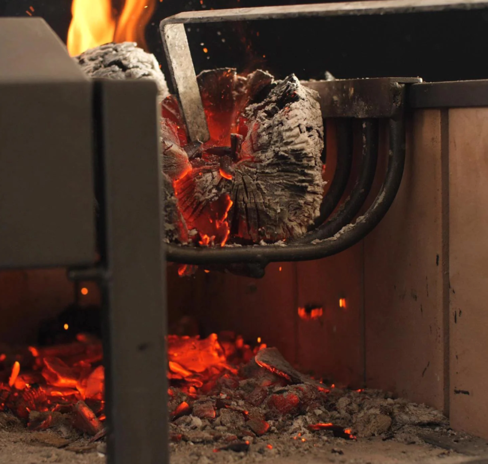 Separate firebox in use on Argentinian style charcoal grill with fire poker stoking coals