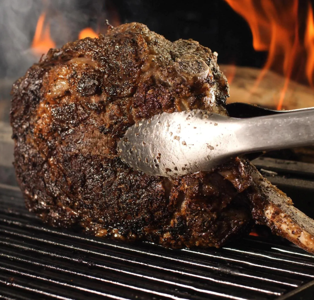 Large tomahawk steak being flipped with metal tongs over charcoal on Argentinian style gaucho grill