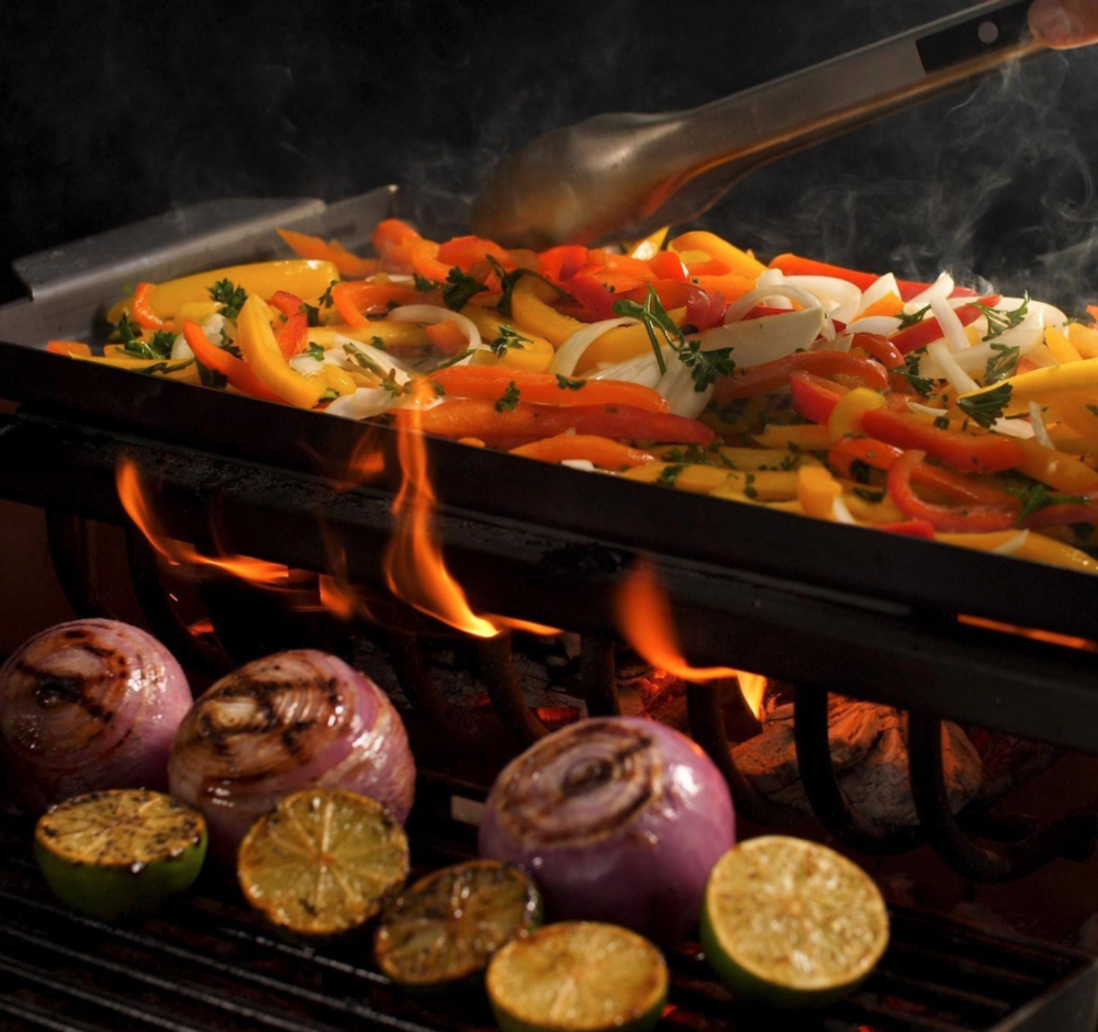 A variety of halved limes and onions being grilled alongside slide yellow, green, orange, and red bell pepper slices on Argentinian style grill